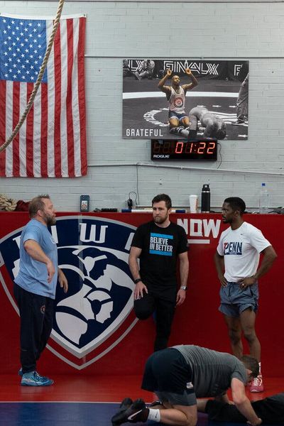 Three men stand beneath an American flag and a large photo of Perry celebrating a victory in a wres…