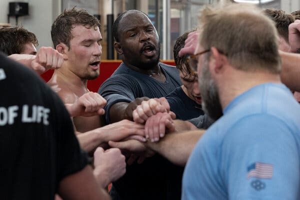 A group of sweaty men that includes Perry stand in a circle while reaching their hands into the cen…