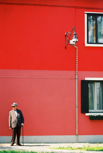 man wearing gray suit jacket and brown dress pants outfit holding cane standing in front of red and…