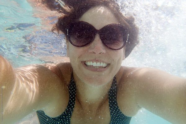 Woman is underwater in a swimming pool wearing sunglasses, smiling for a self portrait