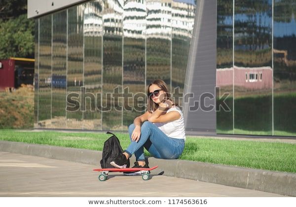 teenage girl sits on a lawn, next to a red skateboard and a backpack.