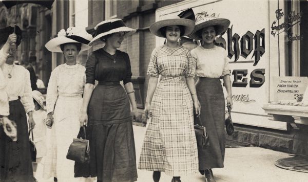 1912 Young women carrying purses and wearing very large hats are walking down a sidewalk. A person …